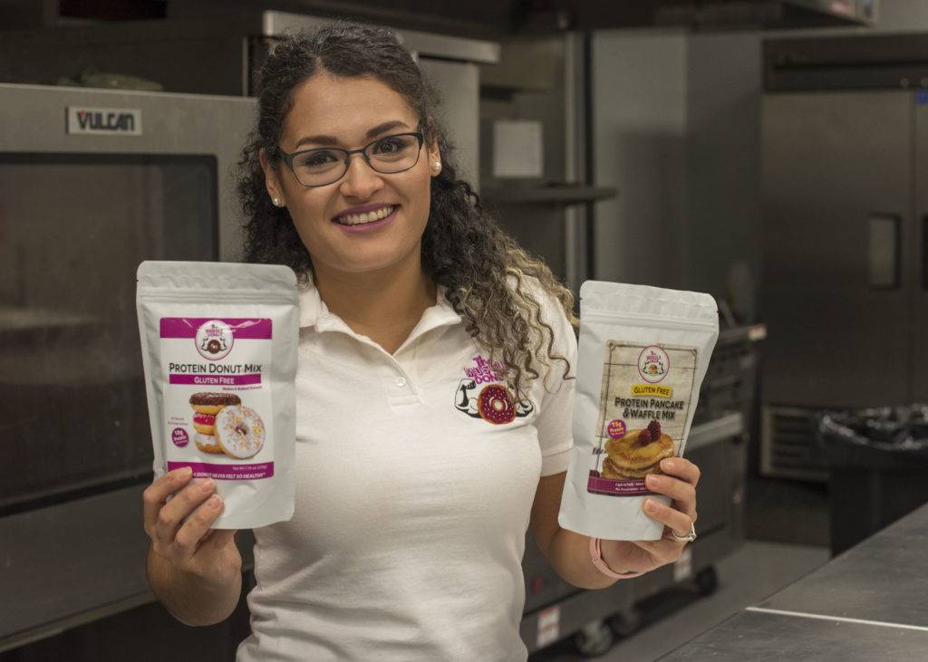 A smiling woman in a kitchen holds two bags of protein mix, one for donuts and one for pancakes