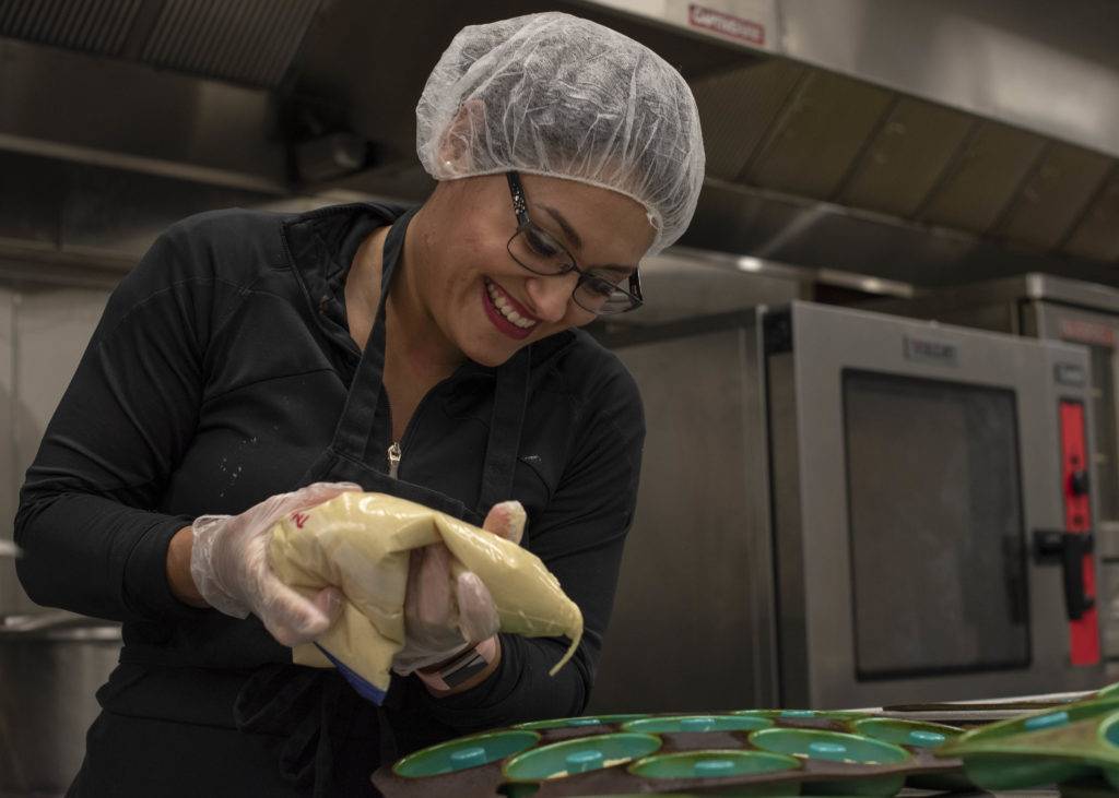 A smiling chef wearing a hairnet and gloves decorates cupcakes in a kitchen