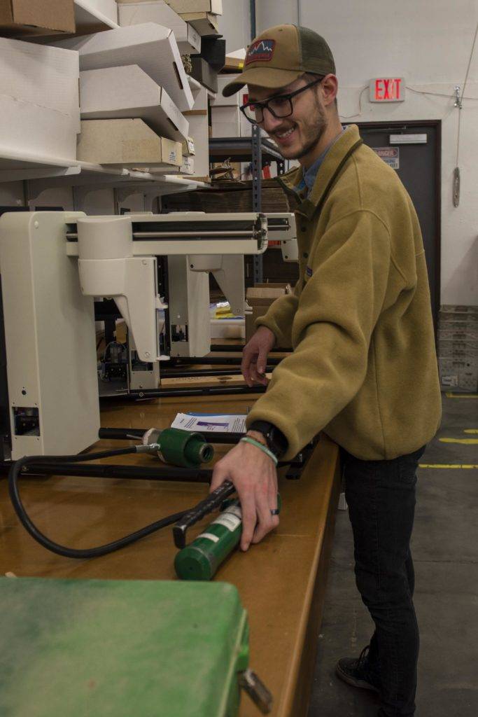 Man in workshop using machine, surrounded by boxes and tools