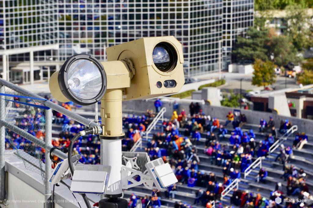 A surveillance camera with a spotlight is mounted on a pole overlooking a crowded stadium