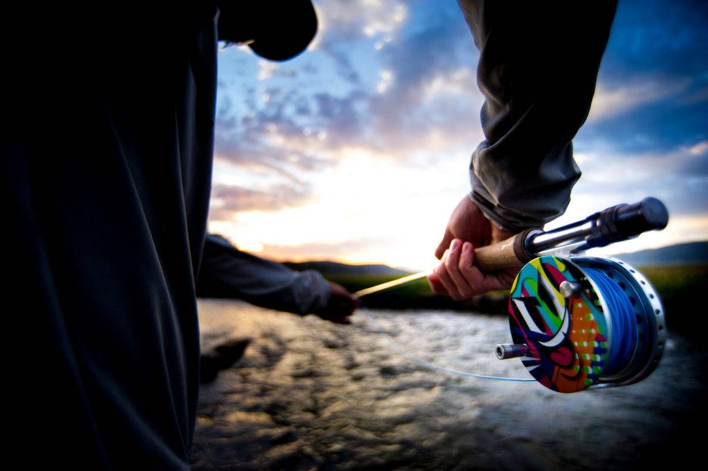 A person's hand holds a colorful fishing reel while casting a line into a river at sunset