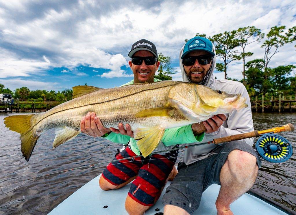Two smiling men proudly hold up a large fish they caught on a sunny day