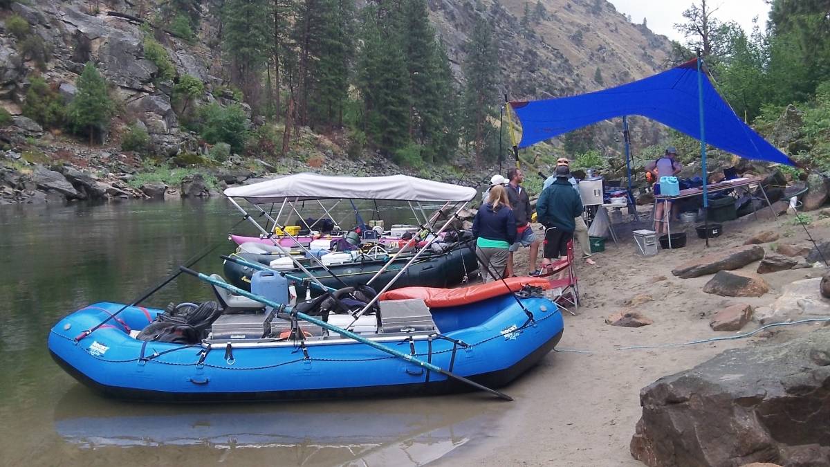 A group of people enjoy a riverside campsite with inflatable rafts and a blue tarp shelter