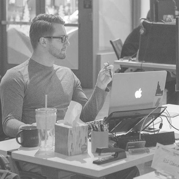 A black and white photo of a man sitting at a desk with a coffee cup, drinking cup, box of tissues, stapler, cup of pens and a laptop.