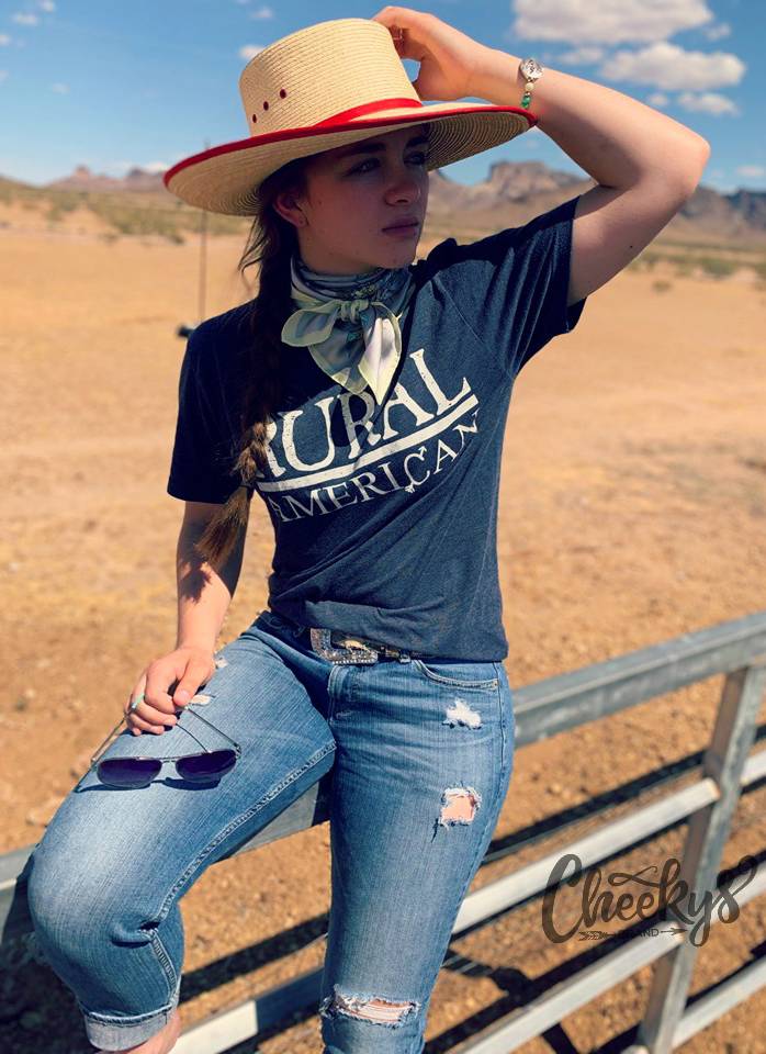 A woman wearing a cowboy hat and a 'Rural American' t-shirt leans on a fence in a desert landscape