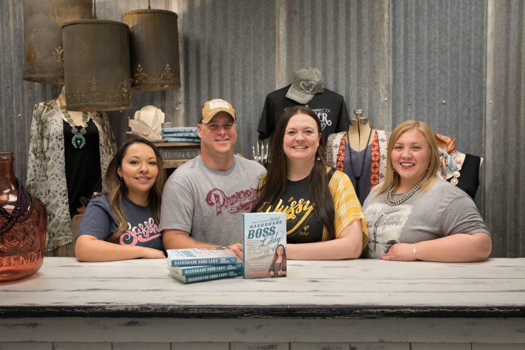 Four smiling people sit at a rustic table in a boutique, surrounded by clothing and accessories