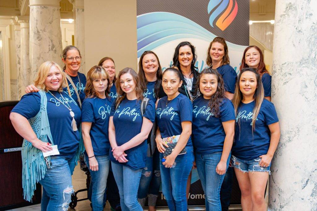 A group of smiling women wearing matching blue shirts pose together indoors