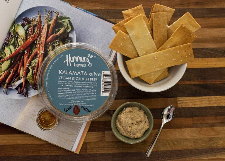 A bowl of vegan and gluten-free hummus with pita chips, a cookbook, and honey on a wooden table