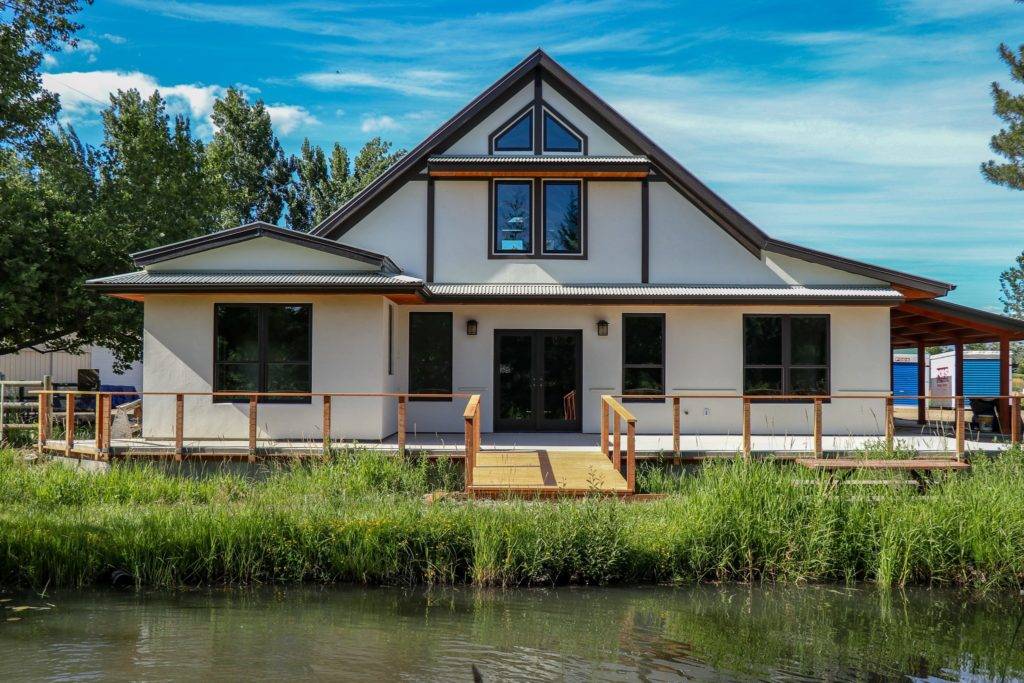 A modern house with a steep roof and large windows overlooks a serene pond surrounded by lush greenery