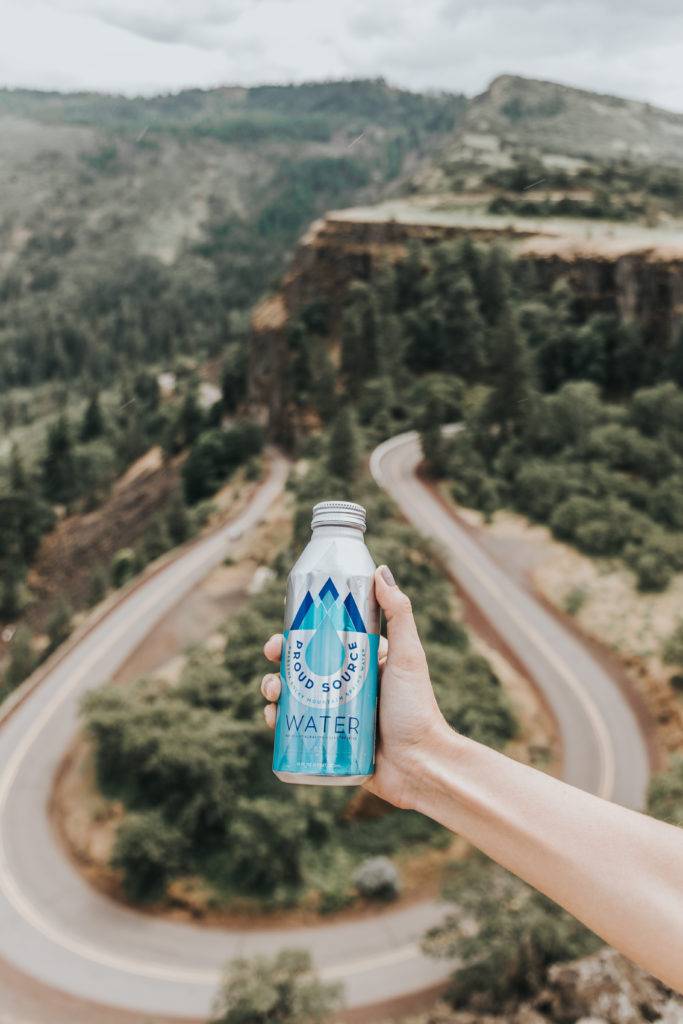A hand holds a bottle of water against a backdrop of a winding mountain road and lush greenery