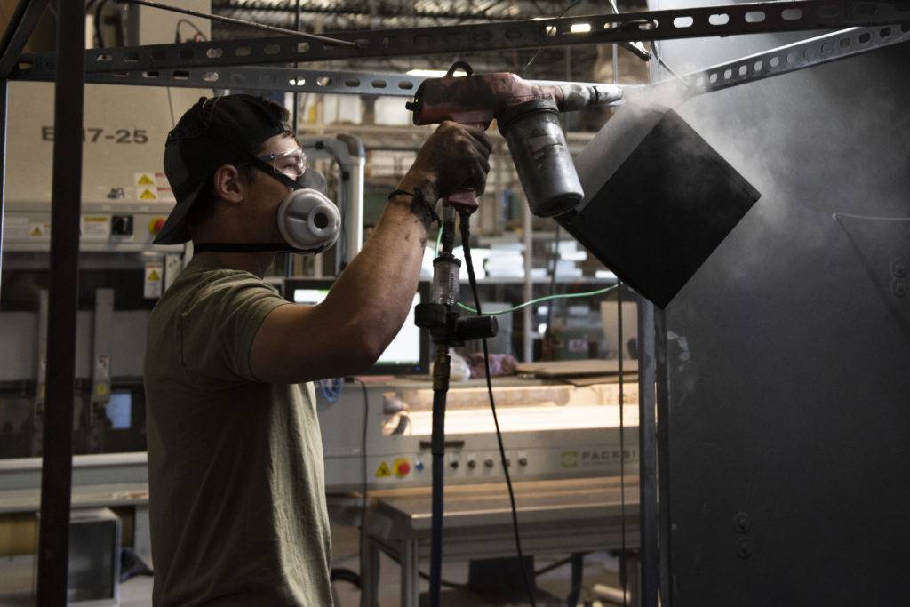 A worker wearing protective gear sprays paint from a handheld device in a factory setting