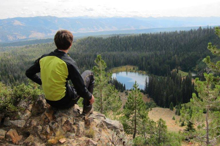 Hiker in yellow jacket sits on rocky peak overlooking serene mountain lake and forest