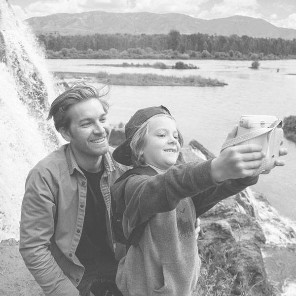 A man and child take a selfie at a waterfall, smiling and enjoying nature