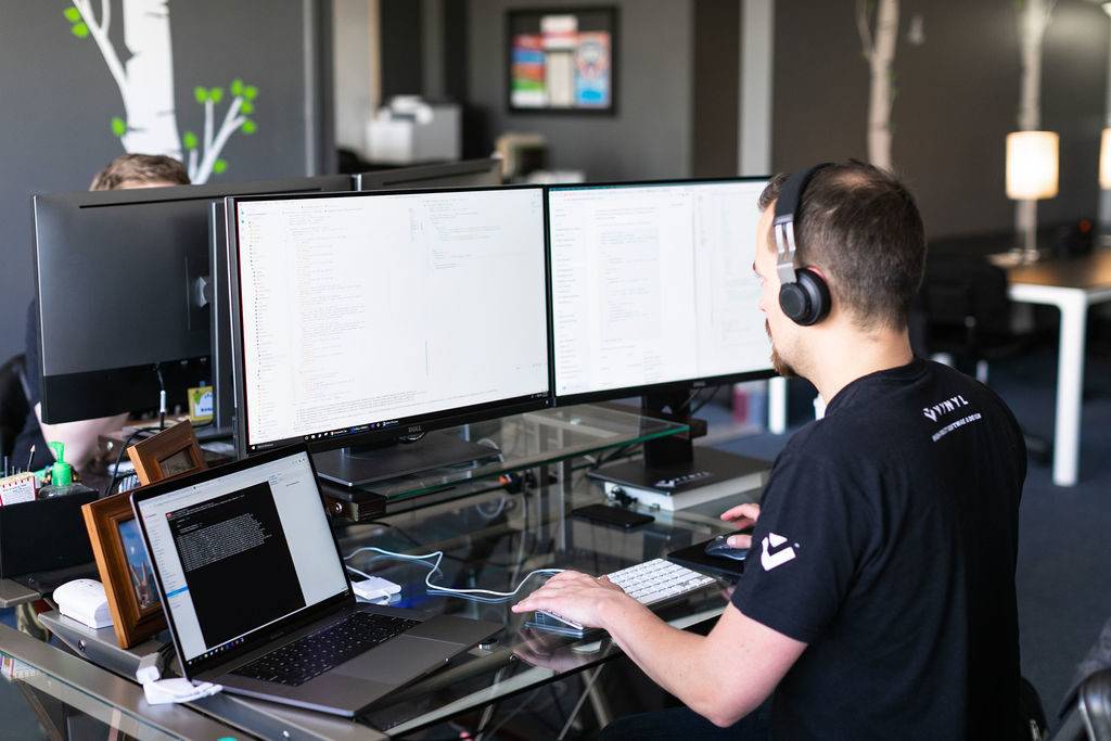 A man wearing headphones is coding on a laptop and two monitors at a desk