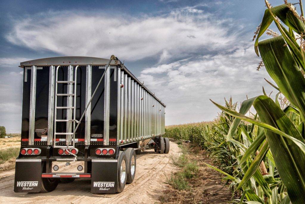 A black semi-truck trailer with a ladder is parked on a dirt road next to a cornfield