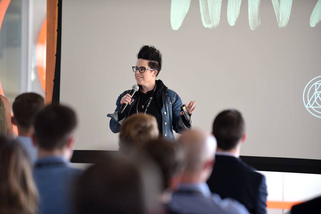 A woman with glasses and a denim jacket speaks into a microphone in front of an audience
