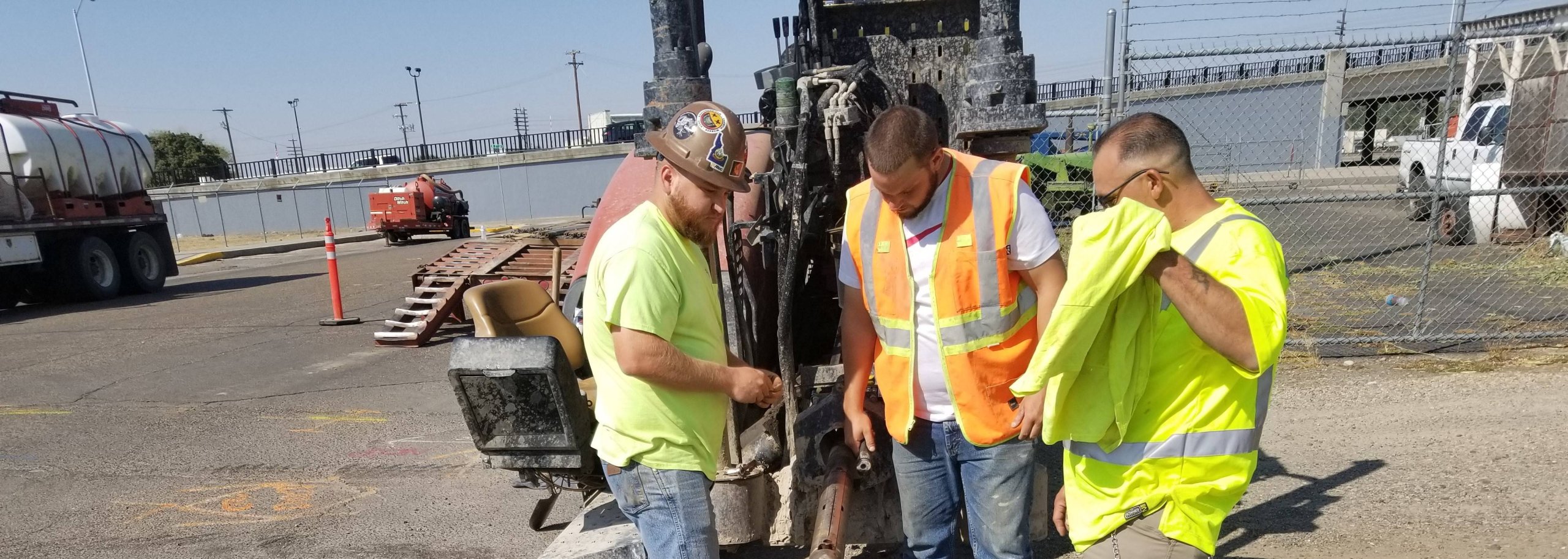 Three workers in high-visibility vests inspect or repair a large industrial machine outdoors