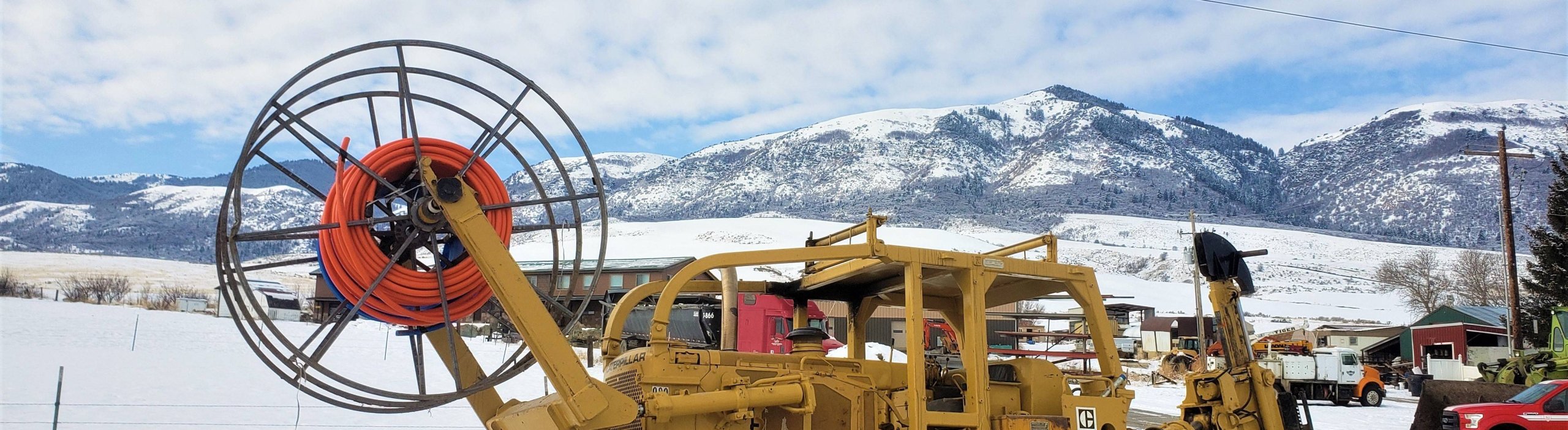 A yellow construction vehicle with a large orange cable spool stands in a snowy field with mountains in the background