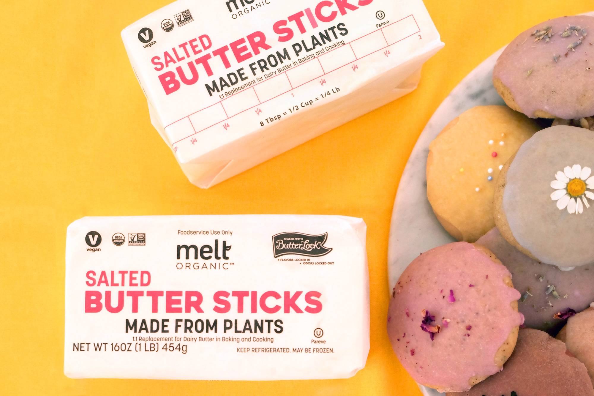 Two packages of Melt Organic Salted Butter Sticks, made from plants, sit on a yellow surface next to a plate of colorful cookies