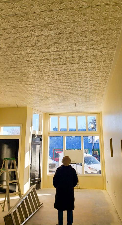 A person in a black coat stands in an empty room with a textured ceiling and large windows