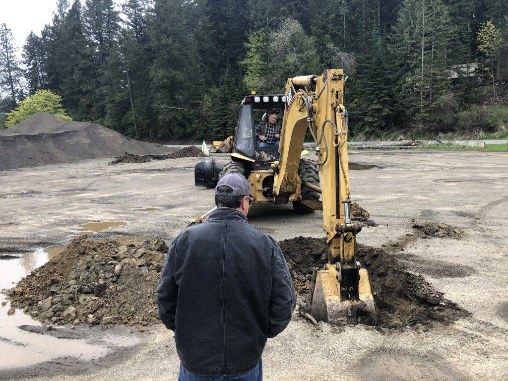 A man observes a yellow excavator digging into a muddy pit surrounded by trees
