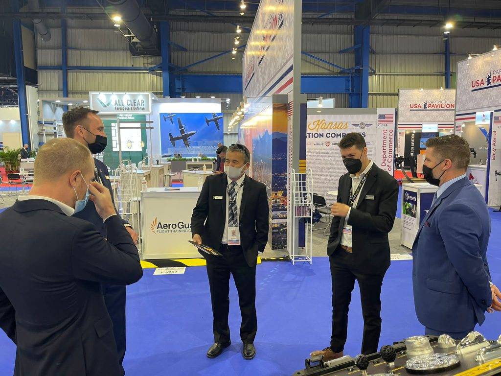 Businessmen in suits and masks converse at an aviation trade show with exhibits and banners
