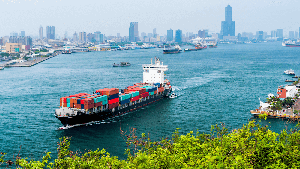 A large container ship with colorful cargo boxes sails through a bustling harbor near a city skyline