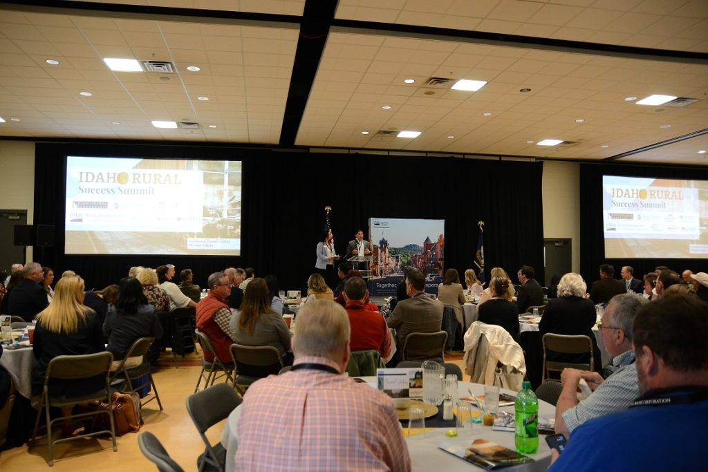 A large conference room filled with attendees at tables, watching a presentation on two large screens