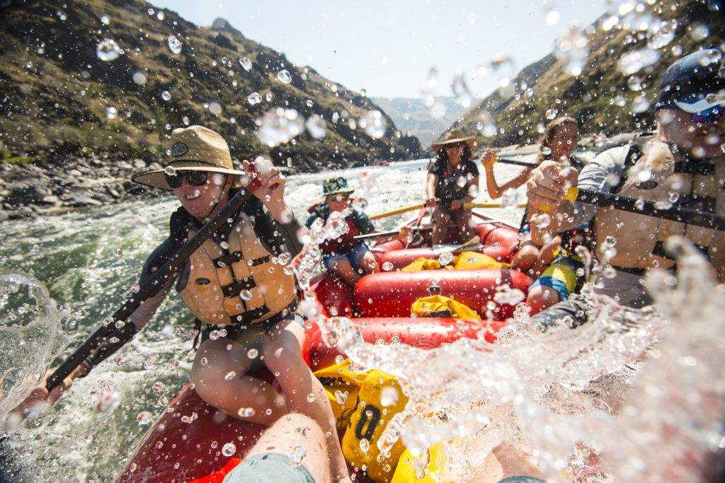 A group of people in a red raft navigate white water rapids, splashing through the river