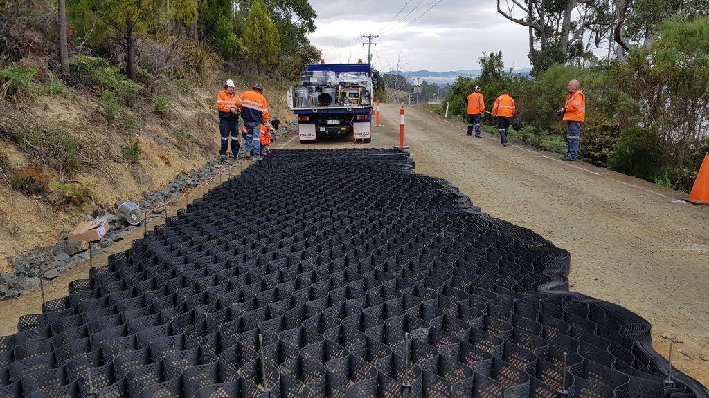 Construction workers in orange vests install a large black honeycomb-like mat on a rural road