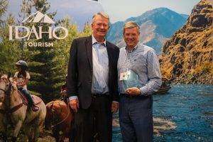 Two men stand in front of an Idaho tourism backdrop with mountains and horses