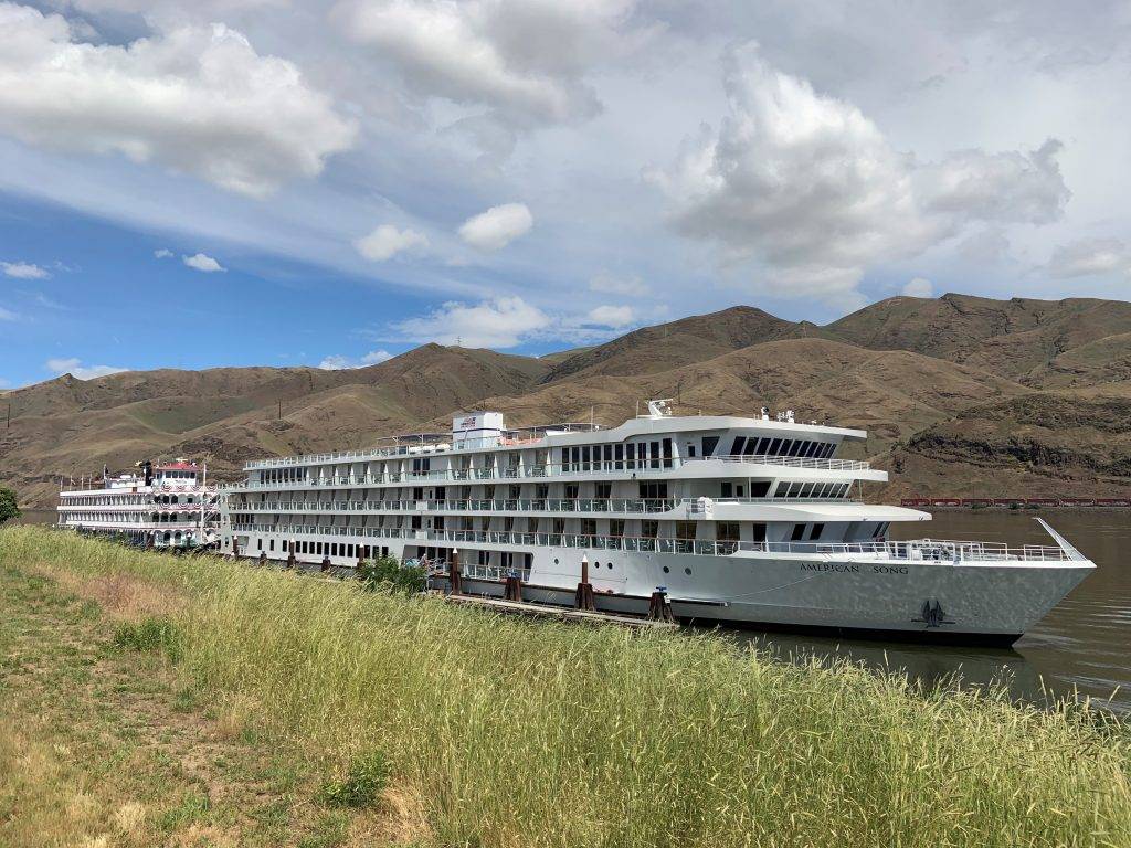 A large white cruise ship is docked at a pier with mountains in the background