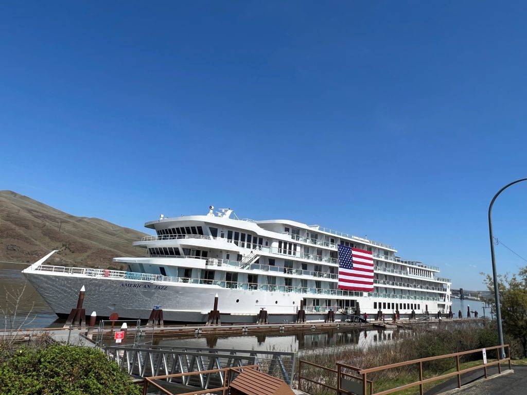 A large white cruise ship with an American flag is docked at a pier with mountains in the background