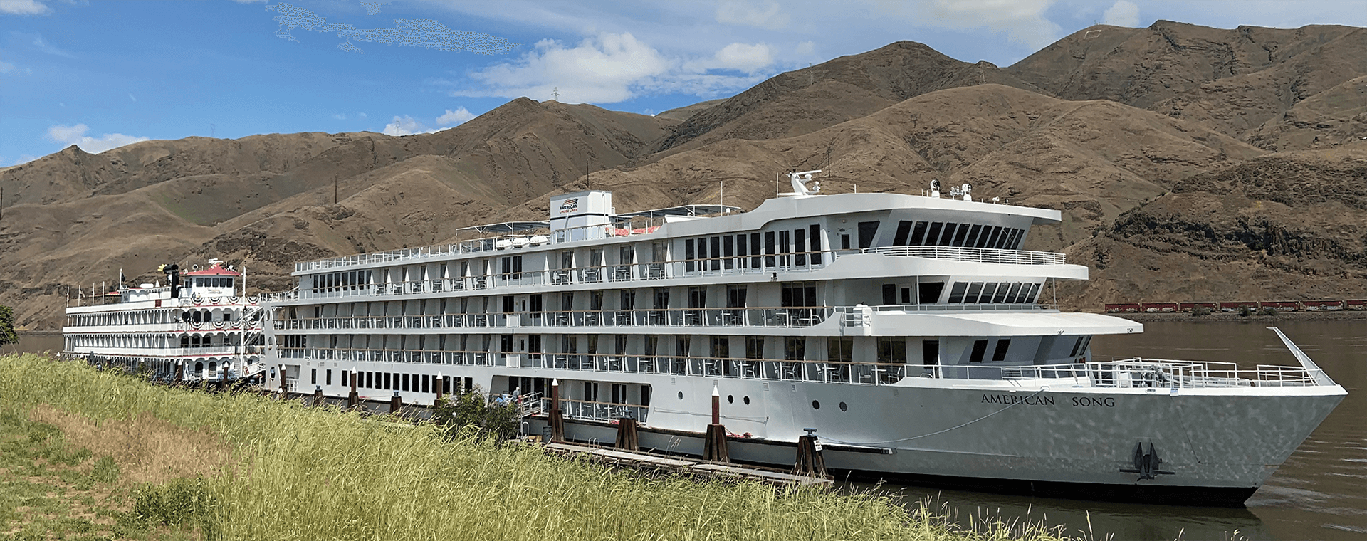 A large white cruise ship is docked at a pier with mountains in the background