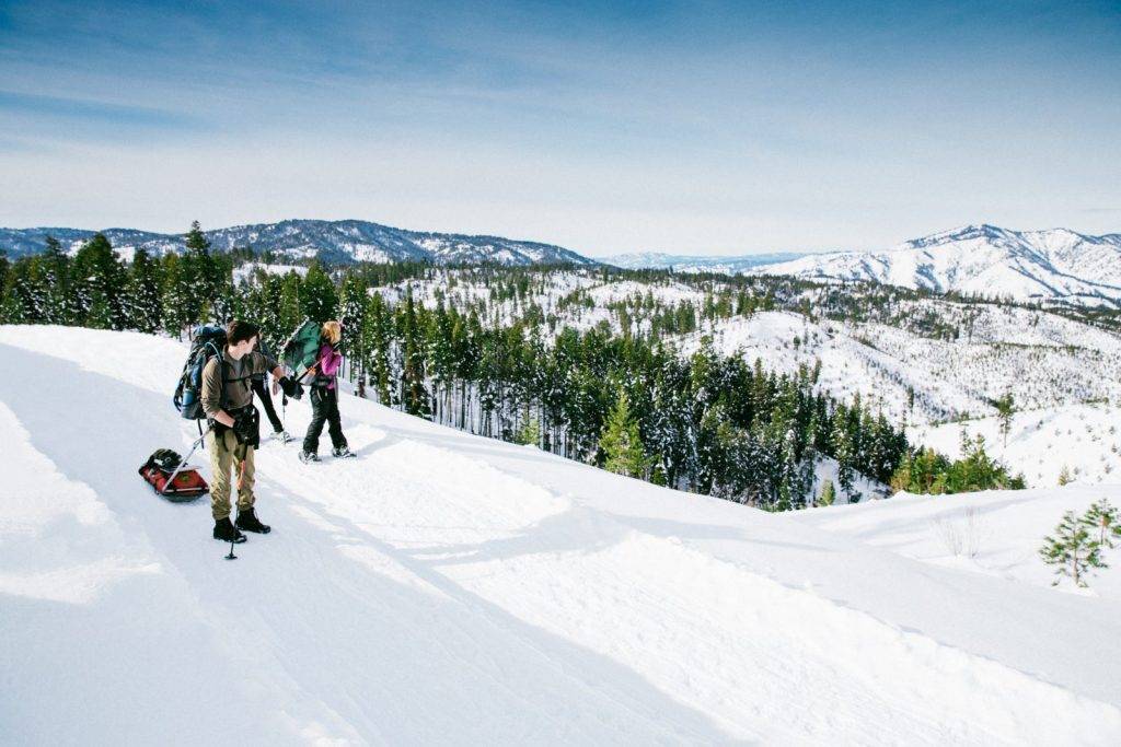 Three hikers with backpacks and snowshoes traverse a snowy mountain slope