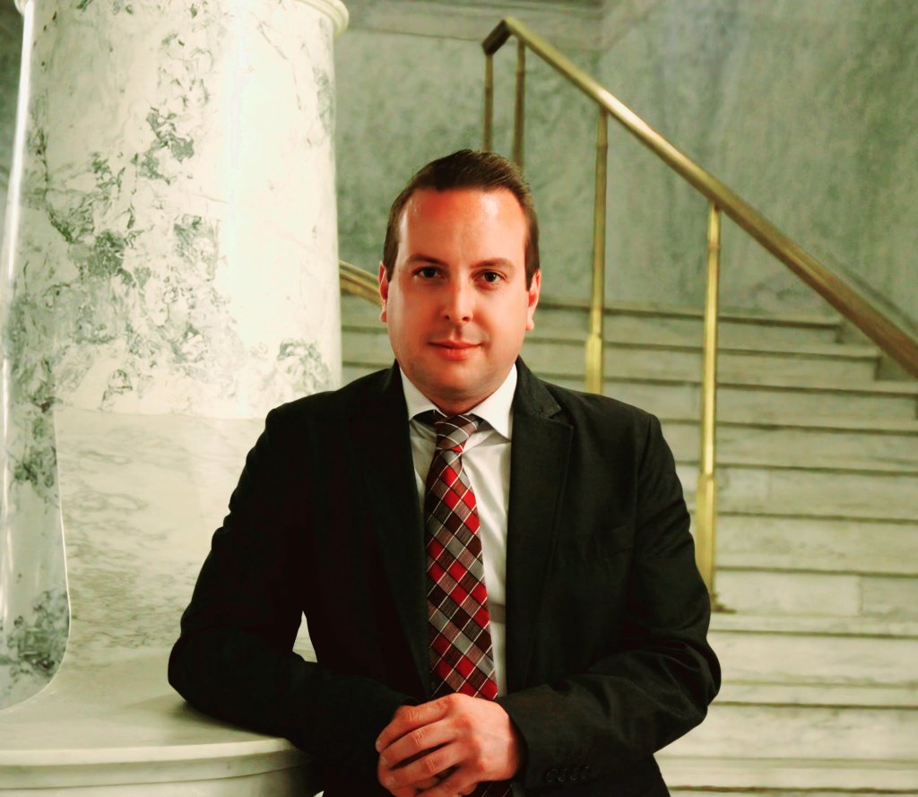 A man in a suit stands at the base of a marble staircase with golden railings