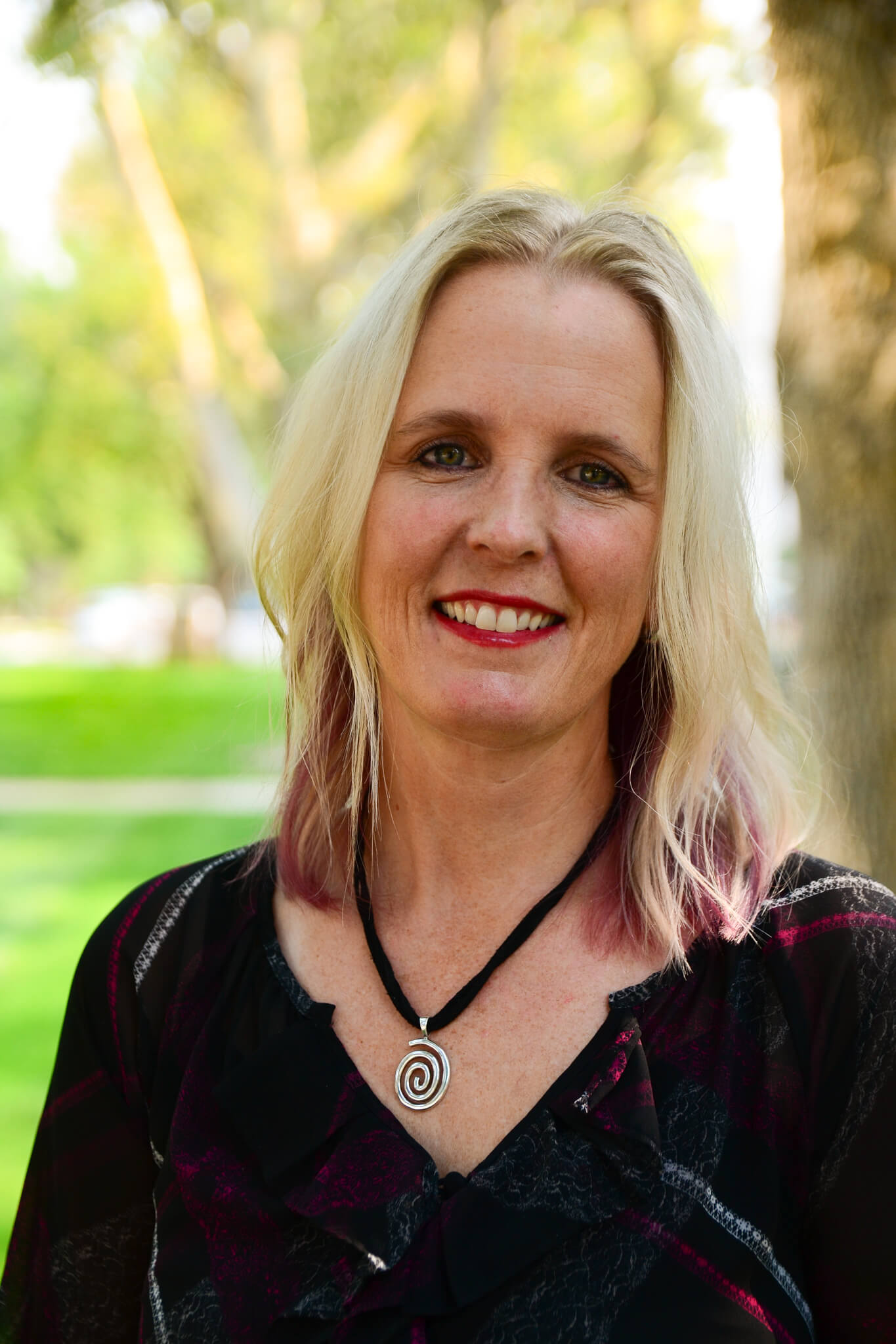 A smiling woman with blonde hair and a spiral necklace stands outdoors