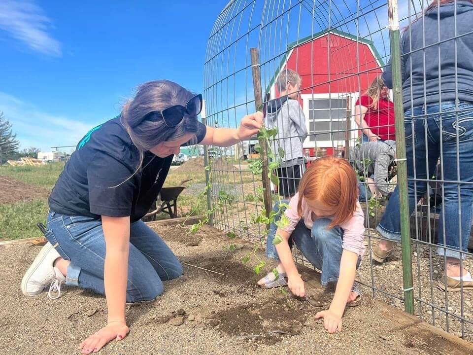 A woman and child kneel in a garden, planting seeds together near a red barn