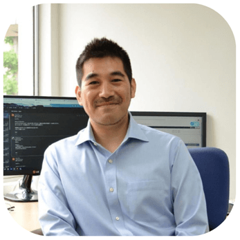 A smiling man sits at a desk with computer monitors displaying code
