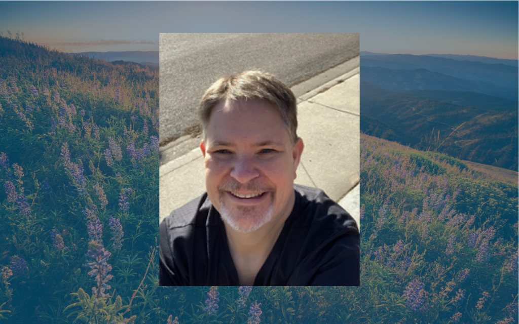 A smiling man stands on a sidewalk surrounded by purple flowers and mountains