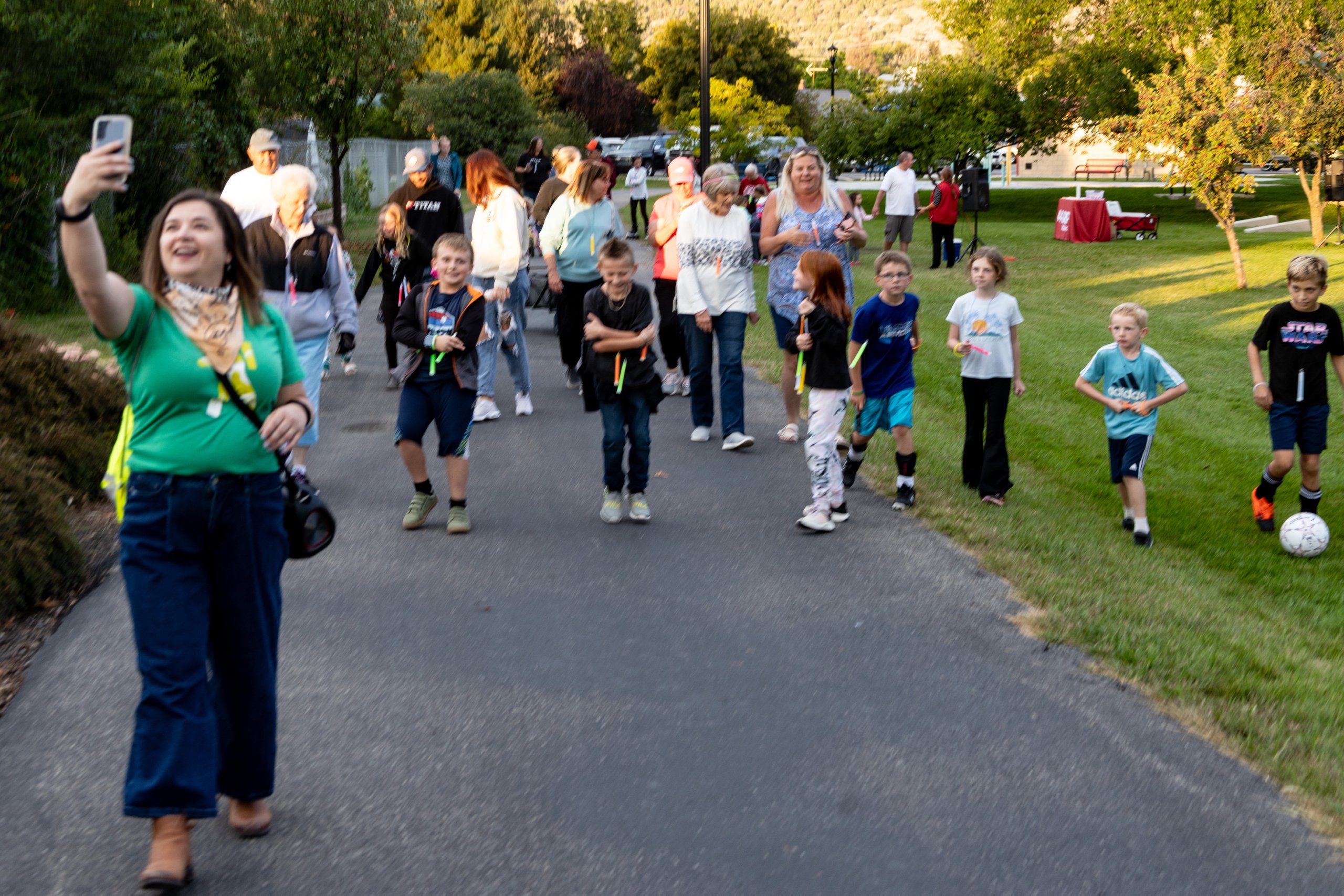 A diverse group of people, including children and adults, walk down a paved path in a park