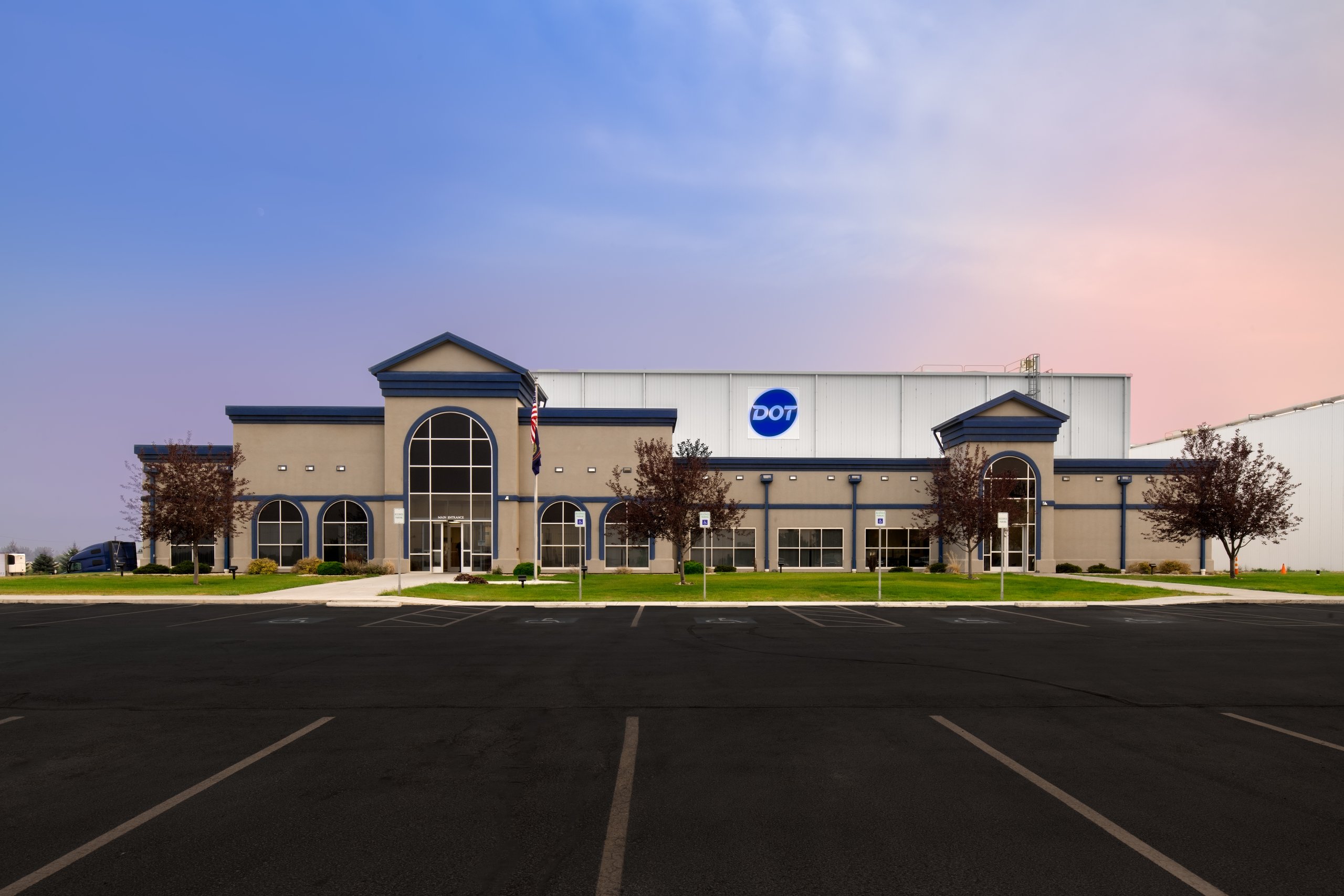 A large industrial building with a prominent DOT logo on the side, set against a colorful sky