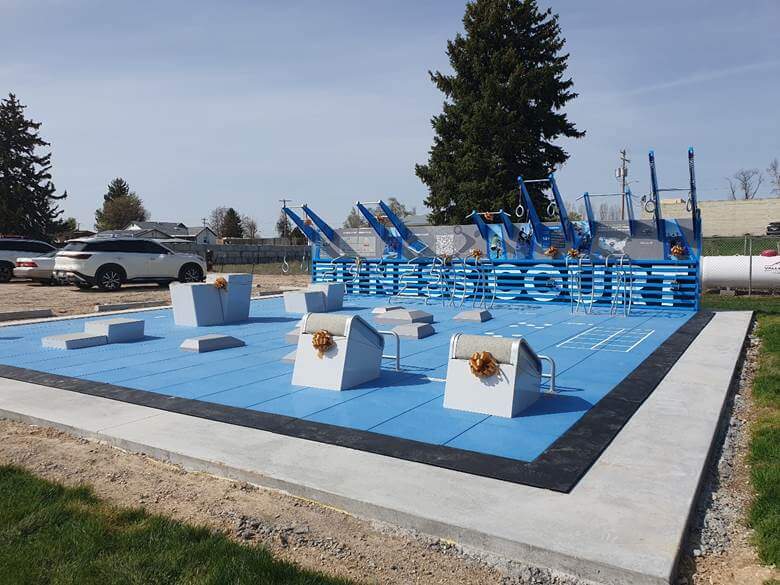 Outdoor fitness court with blue and white exercise equipment, surrounded by grass and trees