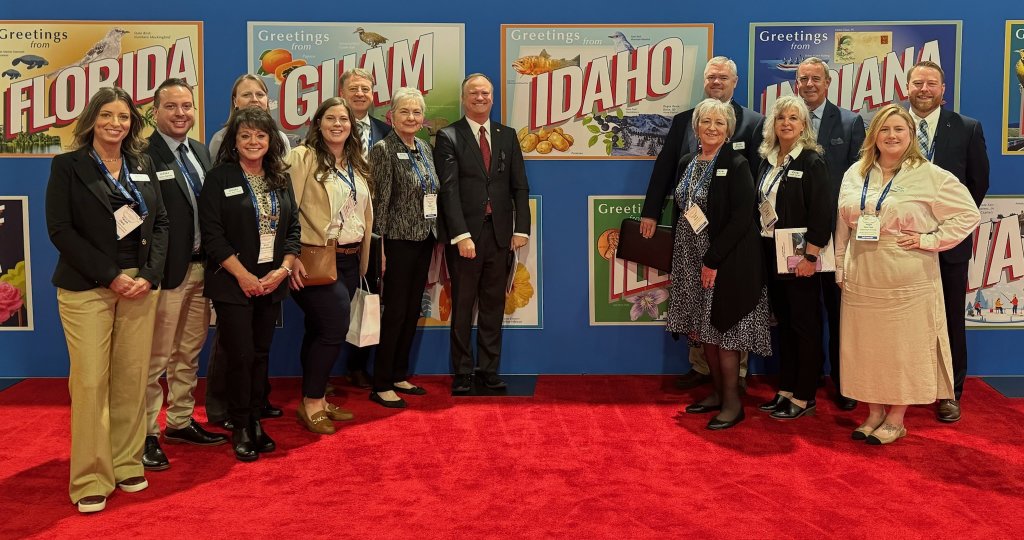 A group of professionals stands on a red carpet in front of colorful state welcome signs