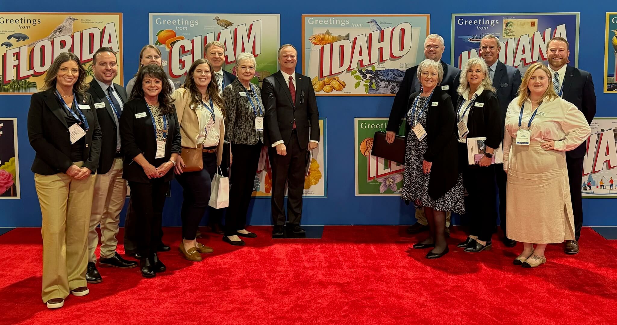 A group of professionals stands on a red carpet in front of colorful state welcome signs