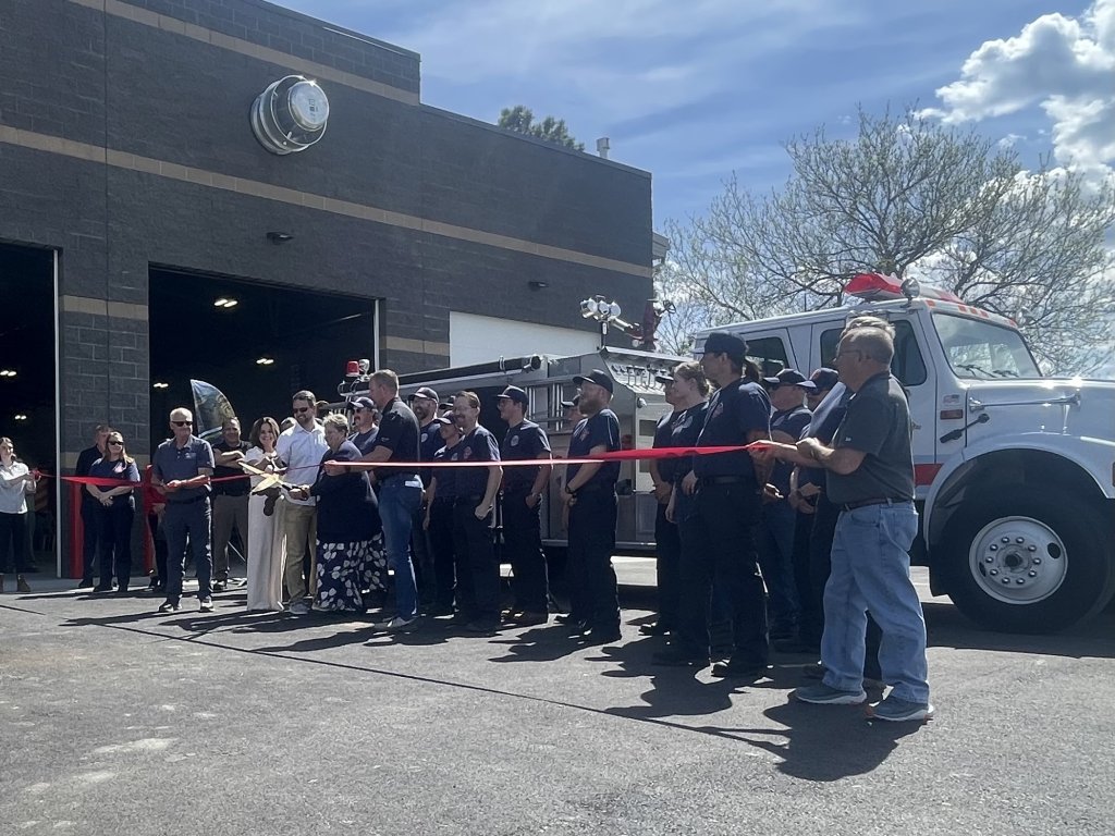 A group of firefighters and officials stand in a line, holding a red ribbon for a ceremonial ribbon-cutting event