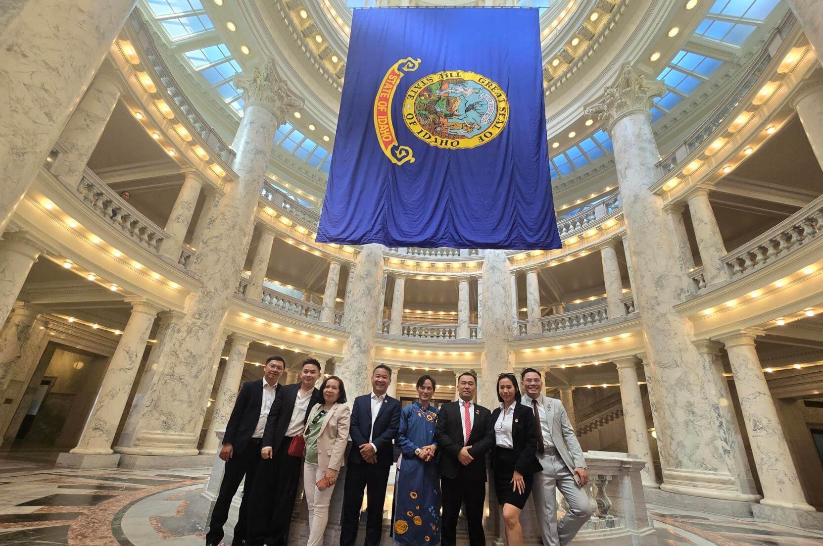 A group of eight people in formal attire stands beneath a large flag in a grand, circular building with marble columns