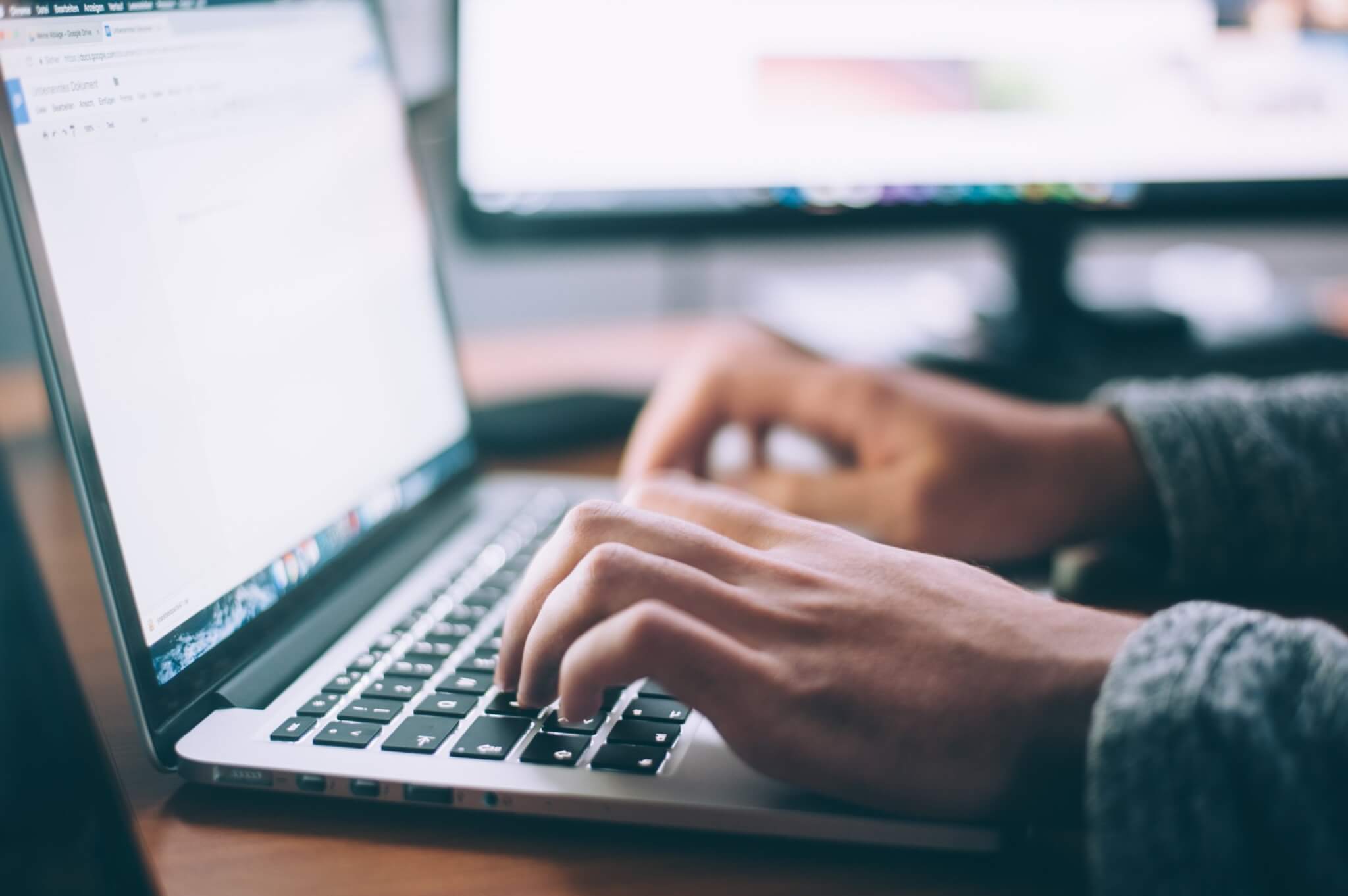 A person's hands are typing on a laptop keyboard with a blurred monitor in the background