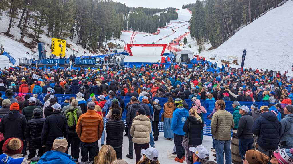 A massive crowd of spectators, dressed in colorful winter gear, gathers at the base of a snowy mountain to watch a ski race, with a large yellow banner and blue fences marking the event area