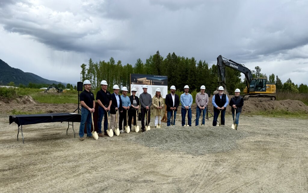 A group of people in hard hats and work clothes stand in a line holding shovels, ready to break ground on a construction site